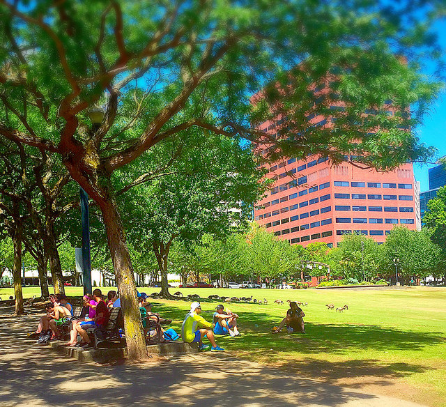People sitting under shade trees in a park with a skyscraper in the background