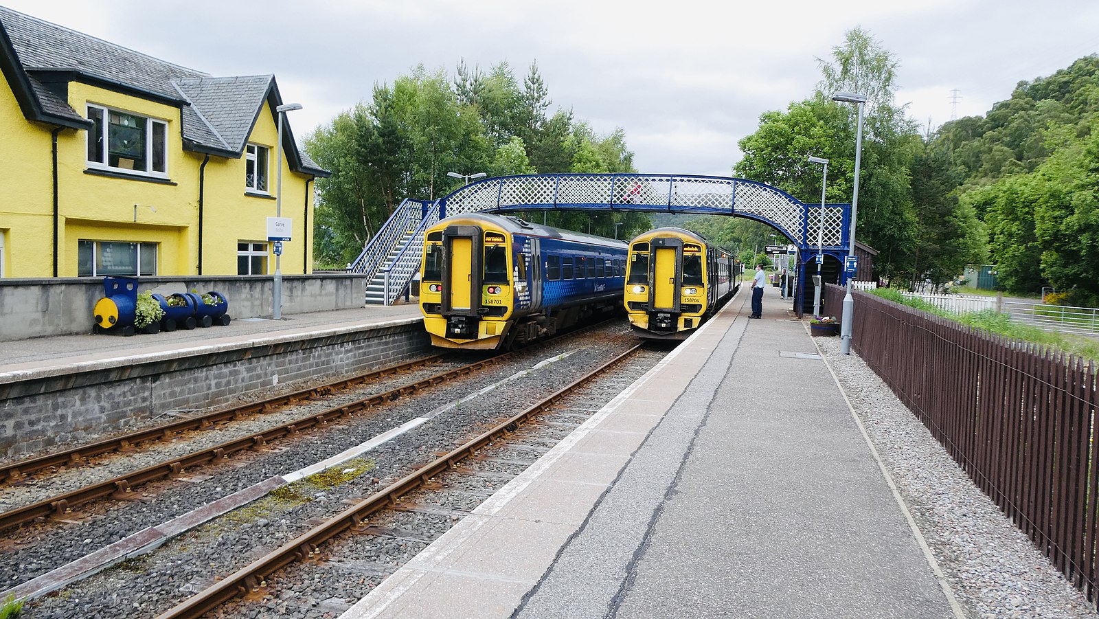 ScotRail trains pull into and out of a station in rural Scotland. There are two tracks with a train on each, with an pedestrian walkway overhead.