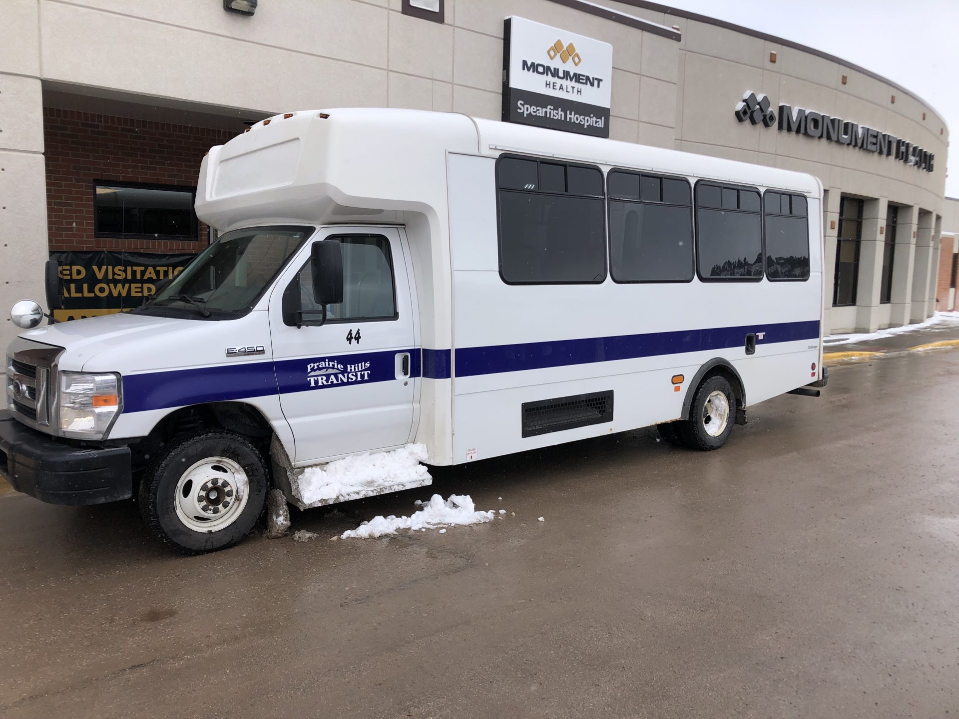 A white bus waits in front of a long hospital building. Snow is on the ground.