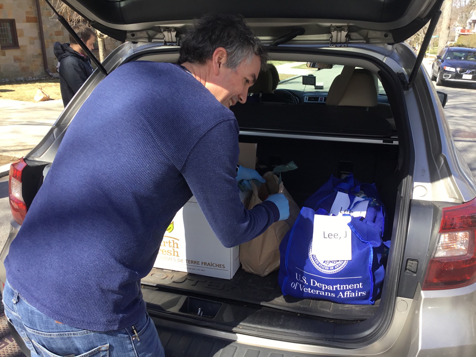 A man with surgical gloves on, half-turns and smiles at the camera as he loads packages into a hatchback's trunk.