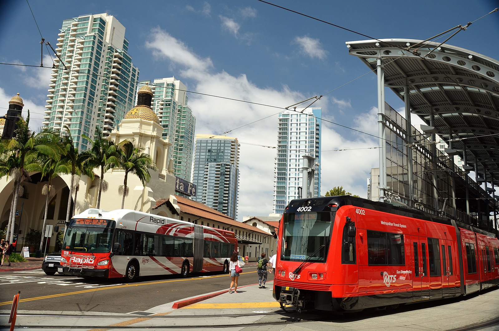 A red train and bus meet at an intersection in San Diego. Glass high rise building and palm trees are in the background.